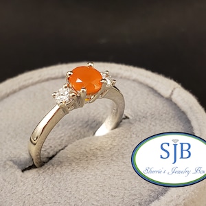 May include: A silver ring with an orange gemstone and two smaller clear gemstones. The ring is in a gray ring box with the text "SJB Sherrie's Jewelry Box".