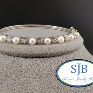 May include: A silver bangle bracelet featuring a row of white pearls and small clear stones. The bracelet is displayed on a grey stand with the text "SJB Sherrie's Jewelry Box".