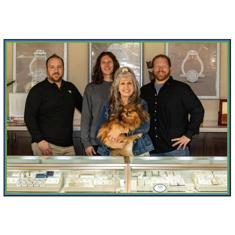 May include: Four people stand in front of a jewelry store display case. Two men and two women are smiling. The woman in the center is holding a small brown dog. The display case is filled with jewelry.