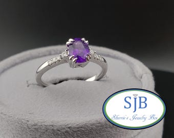 May include: A silver ring featuring a large, oval, deep purple gemstone. Small, clear stones are set along the band. The ring is displayed on a grey velvet surface. The logo "SJB Sherrie's Jewelry Box" is in the lower right.