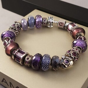 May include: A purple and silver charm bracelet with a variety of beads, including a letter 'C' charm. The bracelet is on a light brown surface.