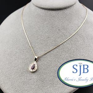 May include: A gold chain necklace featuring a pear-shaped pendant with a dark red gemstone at the centre, surrounded by small clear stones. The logo SJB Sherrie's Jewelry Box is visible in the lower right corner of the image.