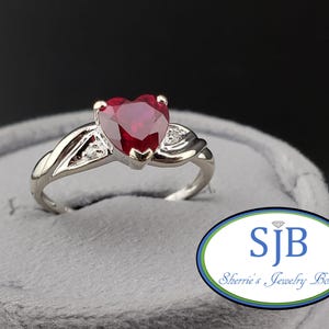 May include: A silver ring with a heart-shaped, deep red gemstone as the centerpiece. The ring has a twisted design on the band and small accent stones on either side of the heart. The ring is displayed on a gray surface.