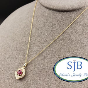 Puede incluir: Un collar de cadena de oro con un colgante en forma de lágrima que presenta una piedra roja rodeada de piedras blancas más pequeñas. El colgante está engastado en un marco de oro. El collar se muestra sobre un fondo gris con un logotipo que dice "SJB Sherrie's Jewelry Box".