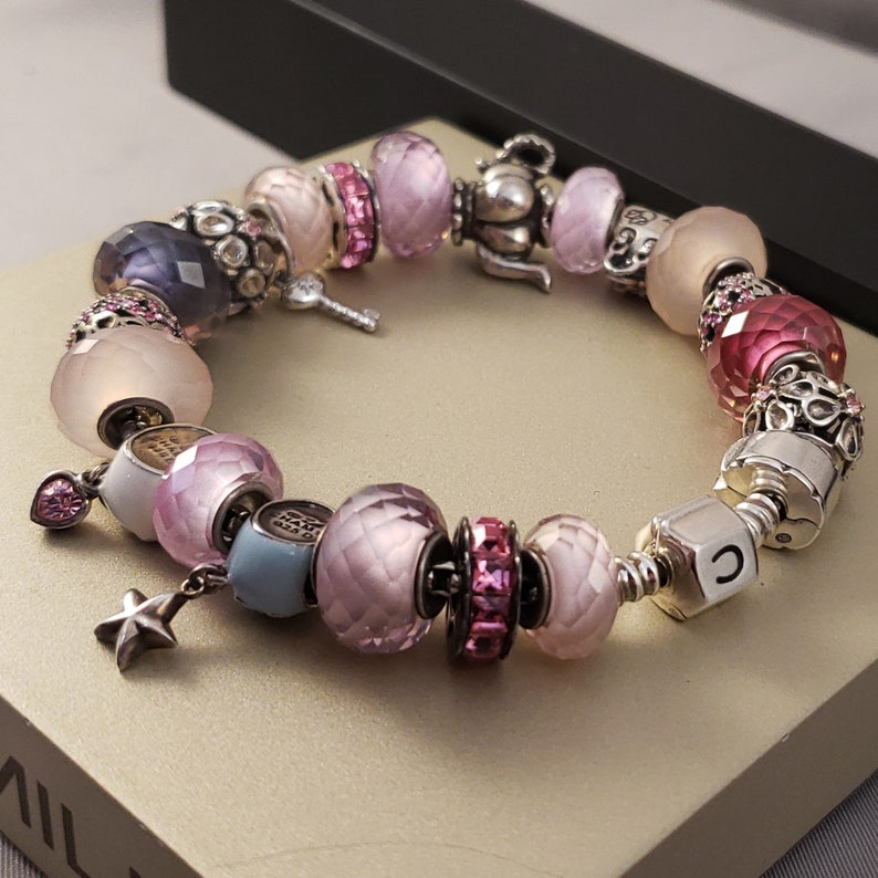 Pu&ograve; includere: Un braccialetto con ciondoli in argento con perline rosa, bianche e blu. Il braccialetto presenta un ciondolo a forma di teiera, una chiave, una stella e una lettera "C".