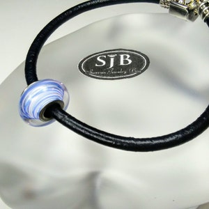 May include: A black leather bracelet with a silver clasp and a blue and white glass bead. The bead has a swirl pattern. The bracelet is on a white surface with a silver tag that says "SJB Jewelry Box".