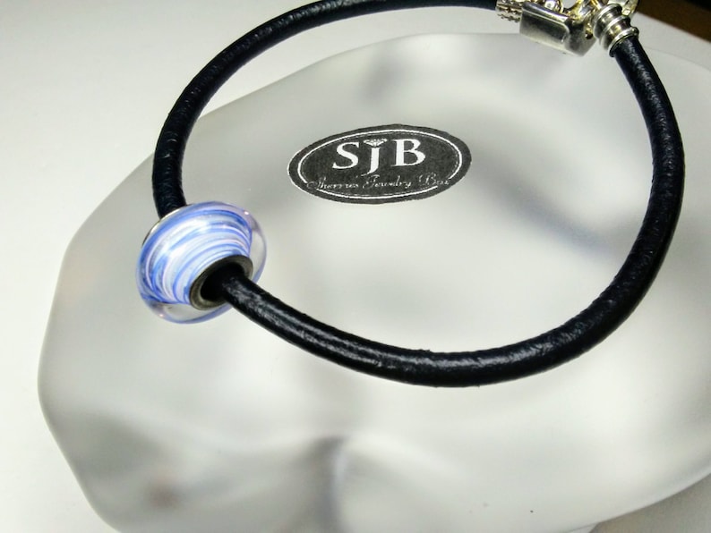 May include: A black leather bracelet with a silver clasp and a blue and white glass bead. The bead has a swirl pattern. The bracelet is on a white surface with a silver tag that says "SJB Jewelry Box".