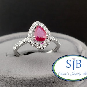Peut inclure: Une bague en argent ornée d'une pierre rouge vif en forme de poire, entourée d'un halo de petits diamants transparents. La monture de la bague est également sertie de petits diamants transparents. La bague est présentée sur un support gris.