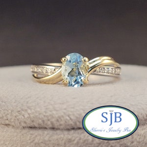 May include: A gold ring with a light blue oval gemstone and a band of small diamonds. The ring is set on a gray background.