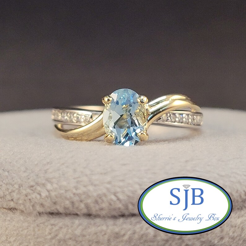 May include: A gold ring with a light blue oval gemstone and a band of small diamonds. The ring is set on a gray background.