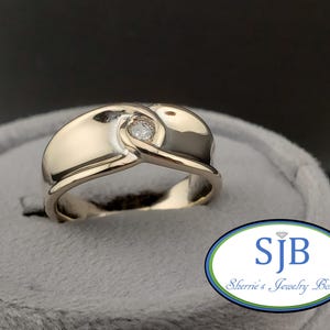 Könnte beinhalten: Ein Goldring mit einem einzelnen Diamanten in der Mitte. Der Ring hat ein einzigartiges Design mit einem gebogenen Band.