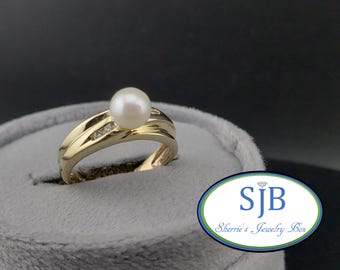 May include: A gold ring with a single pearl and small diamonds set in the band. The ring is displayed on a gray surface with a logo that reads "SJB Sherrie's Jewelry Box".
