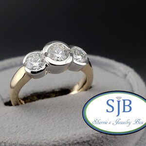 Puede incluir: Un anillo de compromiso de diamantes de tres piedras con una banda de oro amarillo y engastes de oro blanco. La piedra central es la más grande, con dos piedras más pequeñas a cada lado.