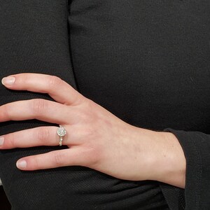 May include: A close-up of a hand wearing a silver ring with a round, clear stone surrounded by smaller stones.