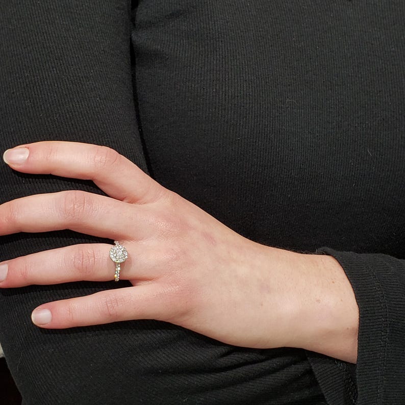 May include: A close-up of a hand wearing a silver ring with a round, clear stone surrounded by smaller stones.