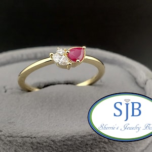May include: A gold ring featuring a pear-shaped ruby gemstone and two smaller white gemstones. The ring is set on a gray velvet surface. The ring is from Sherrie's Jewelry Box.