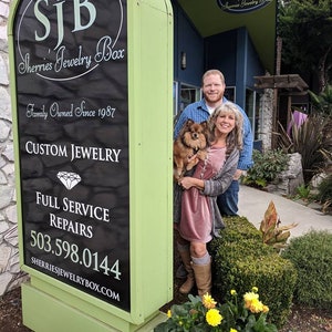 May include: A green sign with black lettering for Sherrie's Jewelry Box, a family owned business since 1987. The sign advertises custom jewelry, full service repairs, and a phone number. A couple and their dog stand in front of the sign.