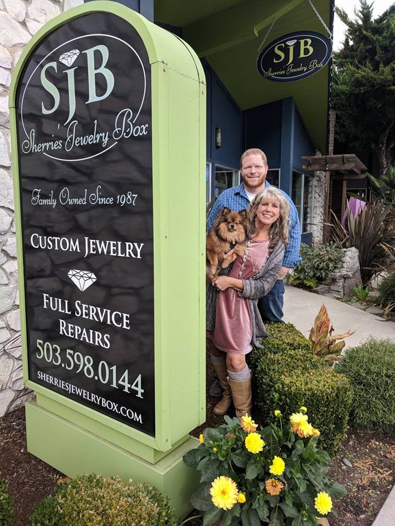 May include: A green sign with black lettering for Sherrie's Jewelry Box, a family owned business since 1987. The sign advertises custom jewelry, full service repairs, and a phone number. A couple and their dog stand in front of the sign.