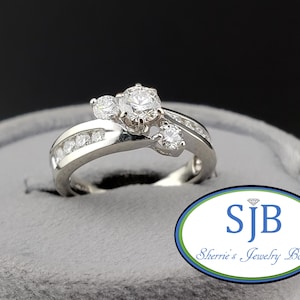 Op de afbeelding: Een zilverkleurige ring met een centrale ronde steen geflankeerd door twee kleinere ronde stenen, met extra kleine steentjes langs de band. De ring wordt gepresenteerd in een grijze fluwelen houder met het logo SJB Sherrie's Jewelry Box.
