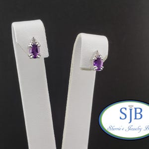 May include: A pair of stud earrings with a silver setting and a rectangular, deep purple gemstone. A small, clear accent stone is set above the purple stone. The earrings are displayed on white stands against a black background.