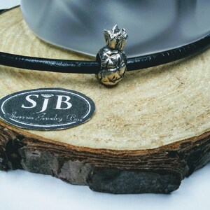 May include: A black leather bracelet with a silver pineapple charm. The bracelet is on a wooden surface with a label that says "SJB Sterling Jewelry Box".