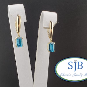 May include: A pair of gold-coloured earrings featuring rectangular, light blue gemstones. The earrings are displayed on white stands against a dark background. The earrings have a lever back closure.