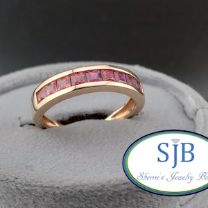 May include: A gold-colored ring with a band of square-cut, bright red gemstones. The ring is set in a gold band and is displayed on a gray surface. The ring is from Sherrie's Jewelry Box.
