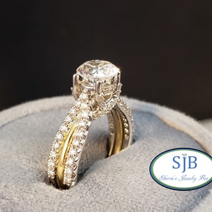 Può includere: Un anello di fidanzamento con diamante, con una pietra centrale rotonda e una doppia fascia. La fascia è incastonata con diamanti più piccoli e ha una sottile striscia dorata.