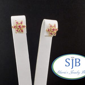Puede incluir: Un par de pendientes de oro en forma de flores de seis pétalos, cada uno con una piedra roja central rodeada por seis piedras rojas más pequeñas en los pétalos. El logotipo SJB Sherrie's Jewelry Box es visible en la esquina inferior derecha.
