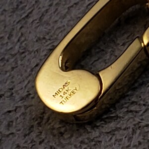 May include: Close-up of a gold-colored metal clasp with the inscription "MIDAS 14K TURKEY".