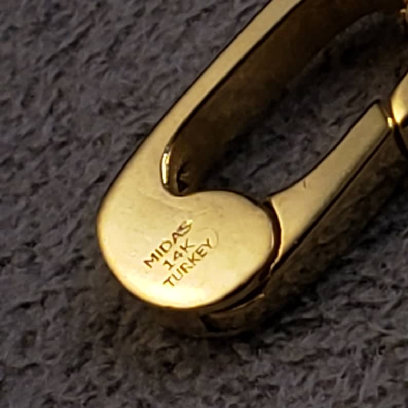 May include: Close-up of a gold-colored metal clasp with the inscription "MIDAS 14K TURKEY".