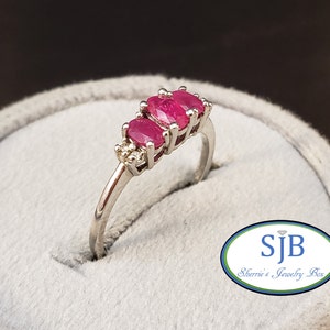 May include: A silver ring with three oval-shaped pink rubies and two small diamonds. The ring is on a gray velvet ring box with the text "SJB Sherrie's Jewelry Box".