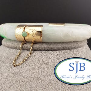 Può includere: Un braccialetto in giada bianca con un design floreale intagliato. Il braccialetto ha una chiusura dorata con un accento di pietra verde. Il braccialetto è su una superficie grigia con un logo che dice "SJB Sherrie's Jewelry Box".