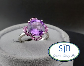 May include: A silver ring with a large, oval-cut purple gemstone, encircled by smaller pink gemstones. The ring is displayed on a grey velvet surface. The ring is a piece of jewellery.