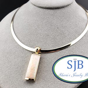 Può includere: Una collana a catena omega in oro con un ciondolo rettangolare con intarsio in opale bianco iridescente. Il ciondolo è incastonato in una cornice in oro lucido. Il testo SJB Sherrie's Jewelry Box con icona diamante è visibile in basso a destra.