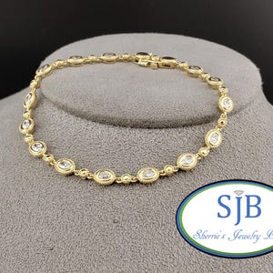 May include: A gold bracelet featuring clear, oval-shaped stones set within a beaded design. The bracelet is displayed on a grey surface. The clasp is visible. The jewellery is from Sherrie's Jewelry Box.