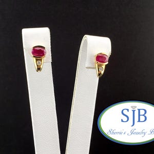May include: A pair of gold earrings featuring an oval-cut deep red ruby at the top and a small round white diamond set below it in a tapered gold setting. The earrings are displayed on white stands with a logo reading SJB Sherrie's Jewelry Box.