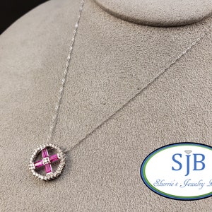 May include: A silver chain necklace with a round pendant featuring a pink cross made of baguette-cut stones surrounded by smaller round stones. The pendant is on a gray background with a logo that reads "SJB Sherrie's Jewelry Box".