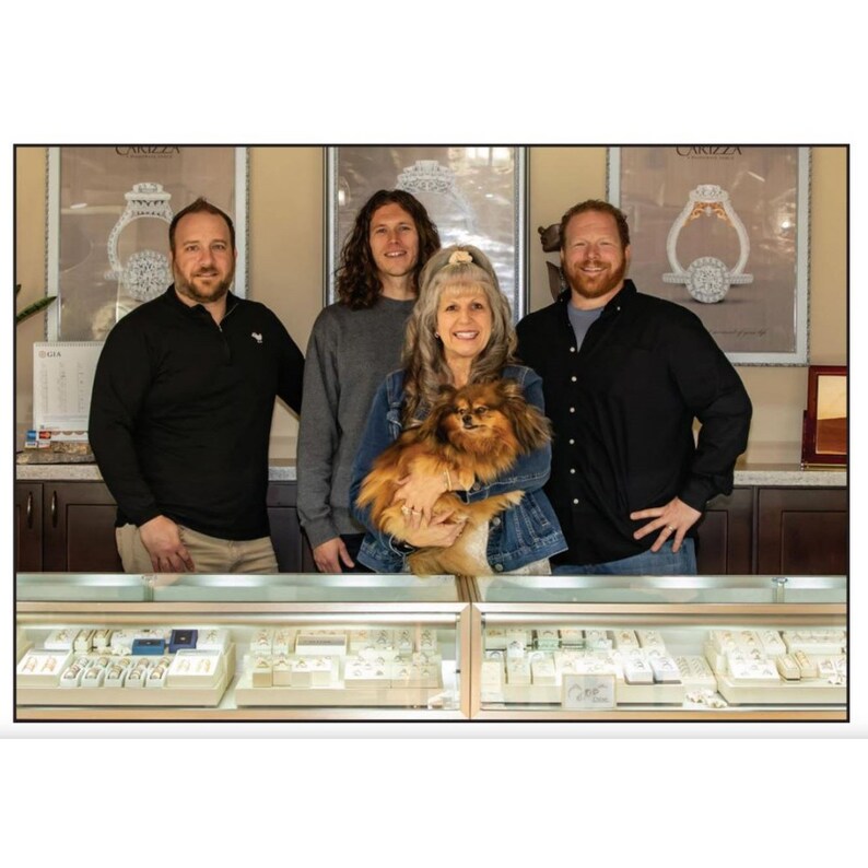 May include: Four people stand in front of a jewelry store display. Two men and two women are smiling. A small brown dog is being held by one of the women. The store has a sign that says "Carizza" and a display of jewelry.