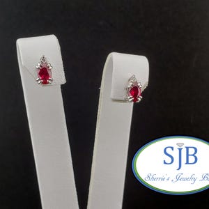 May include: A pair of stud earrings with oval, deep red gemstones set in silver. Each earring has a small, clear accent stone above the ruby. The earrings are displayed on white stands against a dark background.