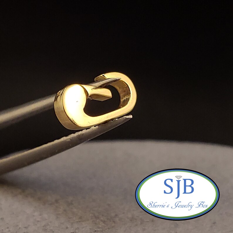 May include: A gold-colored metal clasp for a necklace or bracelet. The clasp is in the shape of a loop with a small bar that fits through the loop to secure it.