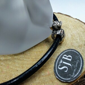 May include: A silver charm with a textured design, resembling a small animal, is attached to a black leather cord. The charm is on a wooden surface with a black and white label that reads "SJB Jewelry Box".