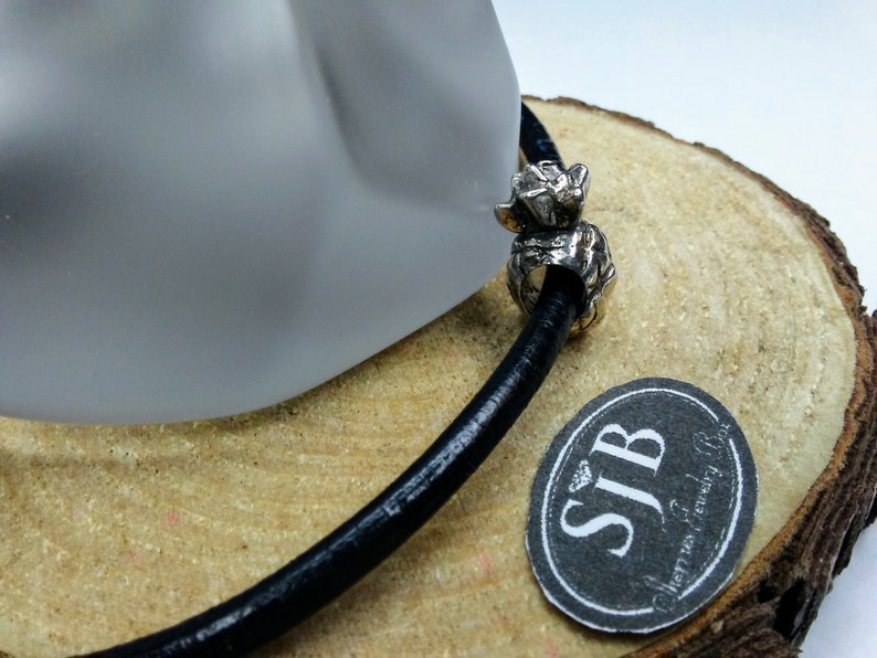 May include: A silver charm with a textured design, resembling a small animal, is attached to a black leather cord. The charm is on a wooden surface with a black and white label that reads "SJB Jewelry Box".