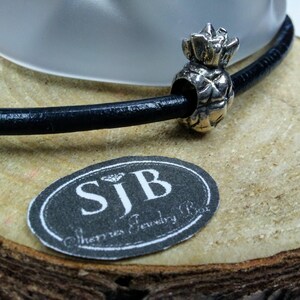 May include: A silver pineapple charm on a black leather cord. The charm is on a wooden surface with a label that reads "SJB Sherri's Jewelry Box".