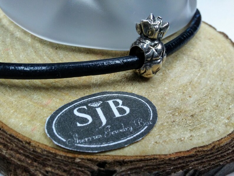 May include: A silver pineapple charm on a black leather cord. The charm is on a wooden surface with a label that reads "SJB Sherri's Jewelry Box".
