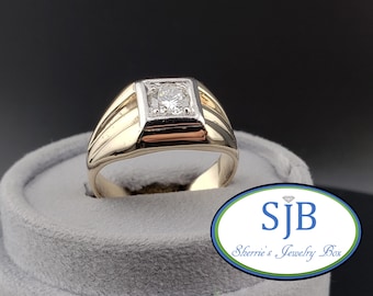 May include: A gold ring featuring a square-shaped setting with a central round brilliant-cut diamond. The band is crafted from polished yellow gold, while the square head holding the stone is finished in a contrasting white metal tone.
