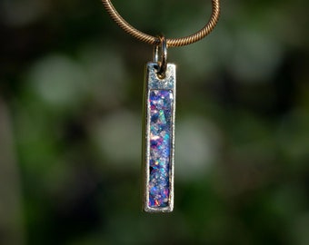 May include: A silver pendant necklace with a rectangular charm filled with iridescent glitter. The charm is suspended from a gold chain.