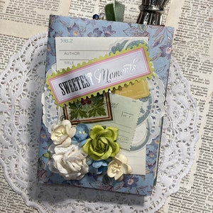 May include: A blue and white scrapbook with a yellow and pink title that reads "Sweetest Moments". The scrapbook is decorated with flowers and a small tag that says "Application".