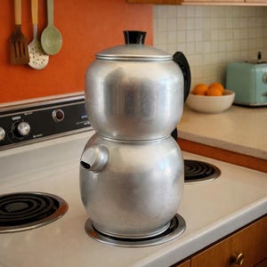 May include: A two-tiered aluminum stovetop kettle with a rounded base and a matching upper section. The kettle features a black handle on the side and a black knob on the lid. It has a short, integrated spout on the lower section and a smooth finish.