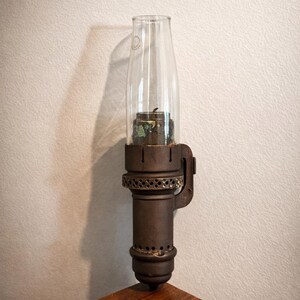 May include: A wall-mounted oil lamp featuring a dark brown, cylindrical metal base with a perforated decorative band and a vertical mounting bracket. A clear, tall glass chimney sits atop the metal burner assembly, which contains a wick.